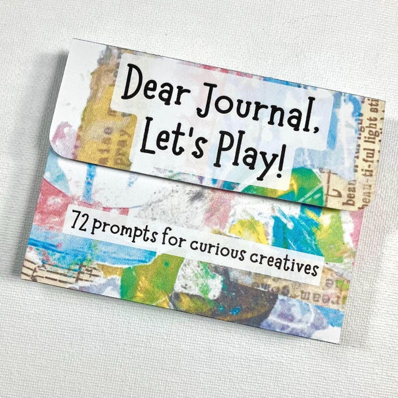 Op de afbeelding: Een journal kaartenset met de tekst "Dear Journal, Let's Play!" en "72 prompts for curious creatives." De kaart heeft een kleurrijk collage-ontwerp met blauwe, gele en roze tinten. De journal set is ontworpen voor creatief schrijven.
