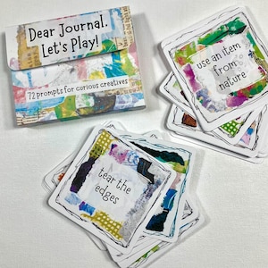 Op de afbeelding: Een set creatieve journaalprompts met een kleurrijk, abstract ontwerp. Op de doos staat "Dear Journal. Let's Play!" en "72 prompts for curious creatives". Afzonderlijke kaarten bevatten prompts zoals "tear the edges" en "use an item from nature".