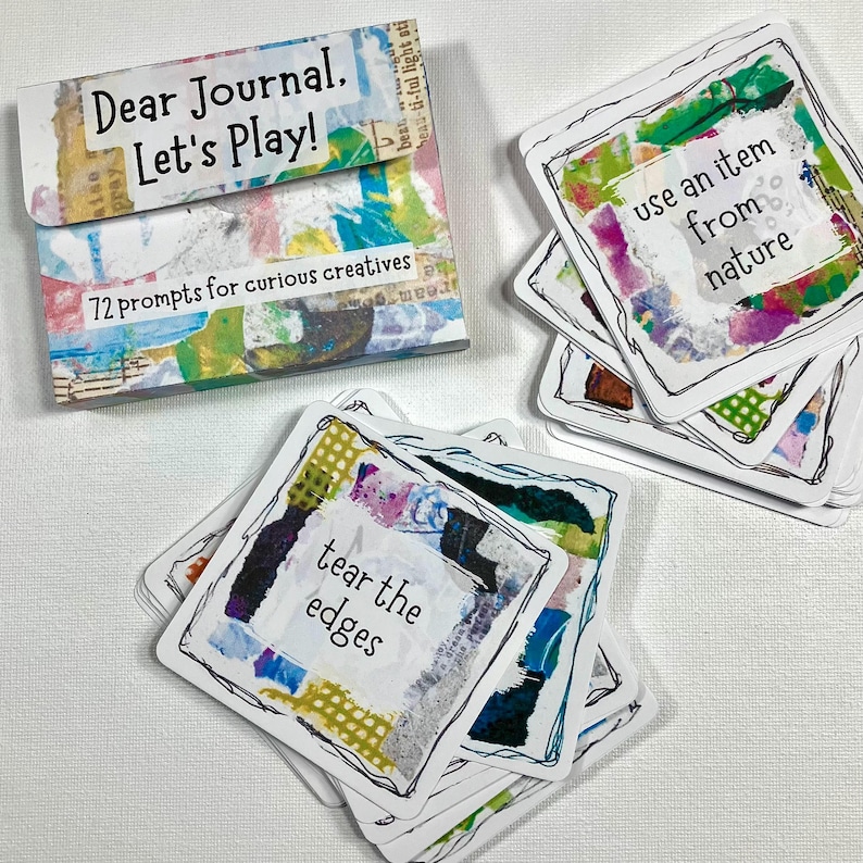 Op de afbeelding: Een set creatieve journaalprompts met een kleurrijk, abstract ontwerp. Op de doos staat "Dear Journal. Let's Play!" en "72 prompts for curious creatives". Afzonderlijke kaarten bevatten prompts zoals "tear the edges" en "use an item from nature".