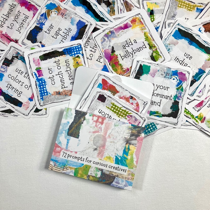 Op de afbeelding: Een verzameling kunstjournaal-promptkaarten met kleurrijke abstracte ontwerpen en handgeschreven tekst. De kaarten zitten in een kleine, versierde envelop met de tekst "72 prompts for curious creatives."