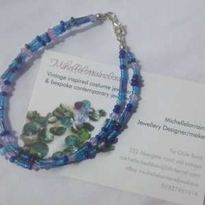 Puede incluir: Un brazalete de cuentas de múltiples hilos con cuentas azules, moradas y plateadas. El brazalete está hecho por Michellelorraine, diseñadora y fabricante de joyas. La tarjeta de presentación incluye información de contacto y el texto "Joyería de época inspirada en el vintage y joyería contemporánea a medida".