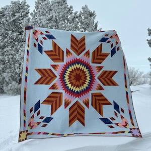 Pode incluir: Um cobertor grande e colorido com um padrão de estrela em forma de explosão em tons de vermelho, laranja, amarelo, rosa, azul e branco. O cobertor é exibido contra um fundo nevado.