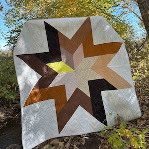 May include: A white quilt with a large, multi-colored star design. The star is made up of brown, tan, and black fabric. The quilt is lying on the ground in a forest setting.