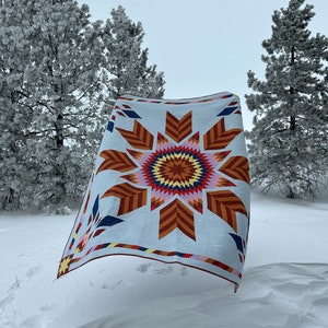 May include: A colorful quilt with a starburst pattern in shades of red, orange, yellow, blue, and white. The quilt is hanging in a snowy landscape with pine trees in the background.