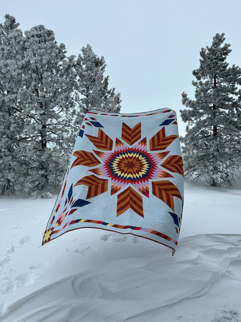 May include: A colorful quilt with a starburst pattern in shades of red, orange, yellow, blue, and white. The quilt is hanging in a snowy landscape with pine trees in the background.