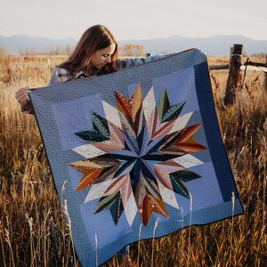 May include: A woman is holding up a blue and brown quilt with a starburst pattern. The quilt is made of various fabrics and has a quilted texture. The woman is standing in a field of long grass.