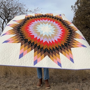 Puede incluir: Una colcha blanca con un gran patrón de estrella en explosión en tonos de marrón, rosa, rojo, morado y azul. La colcha está doblada por la mitad y sostenida por una persona.