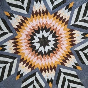 May include: A quilt with a large, multi-colored star in the center. The star is made up of various shades of brown, pink, and yellow. The background of the quilt is a dark blue with a black and white geometric pattern.
