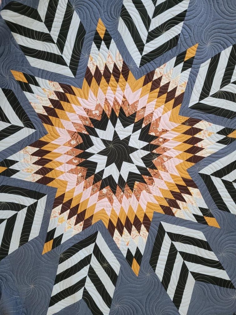 May include: A quilt with a large, multi-colored star in the center. The star is made up of various shades of brown, pink, and yellow. The background of the quilt is a dark blue with a black and white geometric pattern.