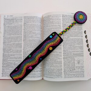 May include: A handmade, black bookmark with a rainbow embroidered design and colorful sequins. The bookmark is placed in an open dictionary. The top of the bookmark has a circular design with a rainbow pattern. The stem is a braided design.
