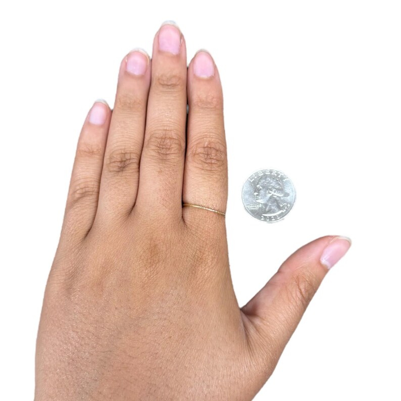 Pu&ograve; includere: Un anello sottile in metallo color oro indossato sul dito medio. L'anello presenta un design a fascia semplice, liscio e sottile. Una moneta statunitense da venticinque centesimi &egrave; posizionata accanto all'anello per fornire una scala visiva.