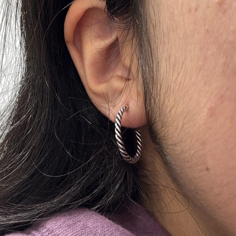 May include: A silver-toned hoop earring featuring a twisted rope-like texture. The circular earring is worn in the earlobe, showcasing a metallic finish with dark oxidized detailing within the grooves of the twisted metal design.
