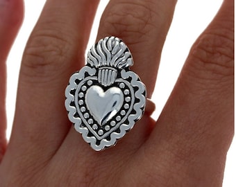 May include: A silver ring with a heart-shaped design. The heart is surrounded by a beaded border and has a flame-like design on top.