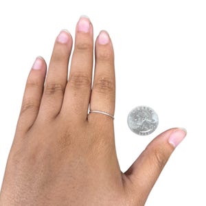 May include: A thin, metallic band ring worn on a finger, positioned next to a United States quarter coin for scale. The ring features a simple, slender design with a smooth, reflective surface, contrasting with the textured detail of the coin.