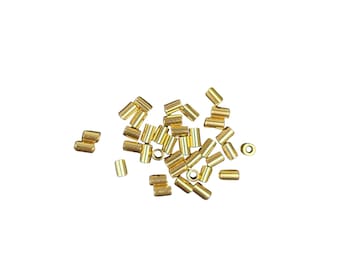 May include: A collection of small, cylindrical, gold-colored metal beads scattered on a white surface. The beads are uniform in size and shape, with a smooth, polished finish. Some beads are lying on their sides, while others stand upright.