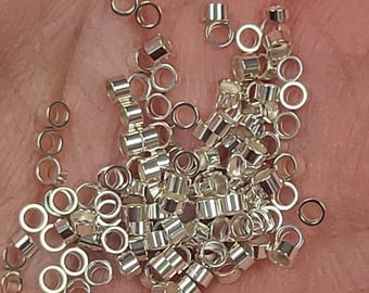 May include: A collection of small, shiny silver metal jewelry findings, including open jump rings and crimp tubes. The findings are scattered across a surface, with a soft, neutral background.