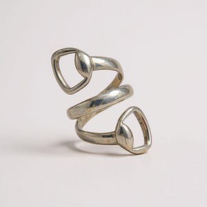 May include: A silver-toned ring with a unique, spiral design. The ring features two open, teardrop-shaped elements at each end of the spiral. The metal has a slightly aged appearance, adding to its vintage appeal. The ring is shown against a plain white background.