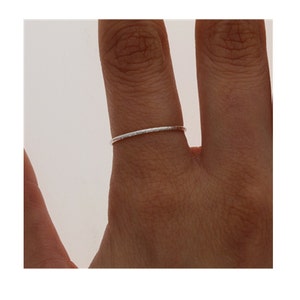 May include: A thin, silver-colored metal ring with a textured, slightly hammered surface. The band is narrow and circular, featuring a reflective finish that catches the light. This simple, minimalist jewelry piece is worn on a finger.