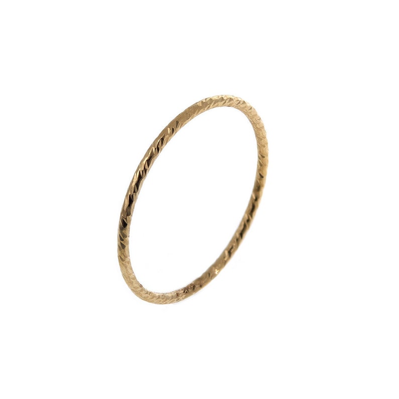 Pu&ograve; includere: Un sottile anello circolare in oro caratterizzato da una superficie testurizzata e sfaccettata che cattura la luce. La fascia presenta una larghezza costante e delicata lungo tutta la circonferenza, creando un gioiello semplice e riflettente.