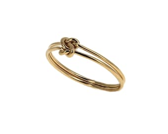 May include: A gold-coloured ring featuring a thin, polished metal band that loops into a small, centred knot design. The band is constructed from a single continuous wire that wraps around itself to form the knot, creating a smooth, reflective surface.