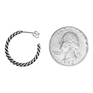 May include: A silver hoop earring with a twisted rope texture and a post back closure, positioned next to a United States quarter coin for scale, which has a diameter of approximately 0.955 inches, showing the relative size of the jewelry piece.