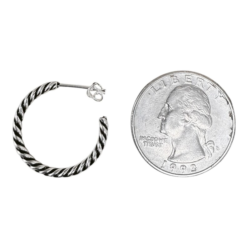 May include: A silver hoop earring with a twisted rope texture and a post back closure, positioned next to a United States quarter coin for scale, which has a diameter of approximately 0.955 inches, showing the relative size of the jewelry piece.