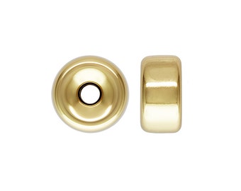 Könnte beinhalten: Zwei goldfarbene Metall-Perlenabstandshalter. Die Perle links hat eine flache Oberseite und ein Loch in der Mitte. Die Perle rechts ist zylindrisch.