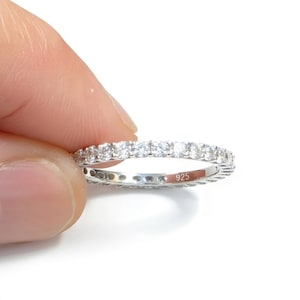 May include: A silver-toned eternity band ring featuring a continuous row of small, clear, round-cut stones set along the outer circumference. The interior of the metal band is clearly engraved with the hallmark text 925, indicating sterling silver.
