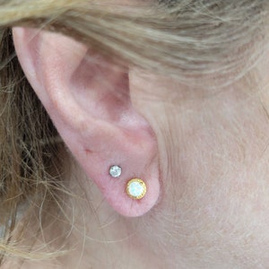 May include: A close-up of a person's ear with two earrings. The earring on the lower lobe is a gold-toned round earring with a white stone in the center. The earring on the upper lobe is a silver-toned stud earring with a small clear stone.