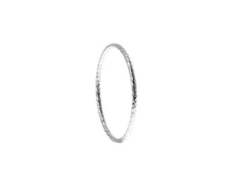 May include: A thin, circular silver ring featuring a textured, faceted surface that reflects light. The band has a consistent, slender profile and a metallic, reflective finish, creating a simple, continuous loop design without any additional stones or settings.