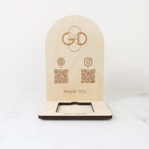 May include: A light-coloured wooden display stand with an arched top. The stand features a logo, QR codes for a website and Instagram, and the words "THANK YOU" and "TAP HERE".