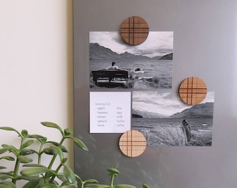 May include: A gray metal surface with three wooden magnets with a plaid pattern. Two black and white photos are attached to the surface with the magnets. One photo shows a person sitting on a bench overlooking a body of water. The other photo shows a person standing in a field of tall grass. A handwritten grocery list is attached to the surface with a magnet. The list includes the following items: apples, bananas, lemons, spinach, onion, feta, eggs, milk, butter, coffee.