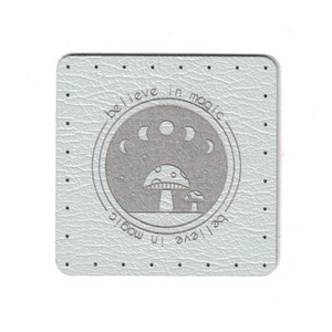 May include: Square, light blue coaster with a textured surface and a dotted border. The center features a gray circular design with a mushroom and moon phases, and the words "believe in magic".