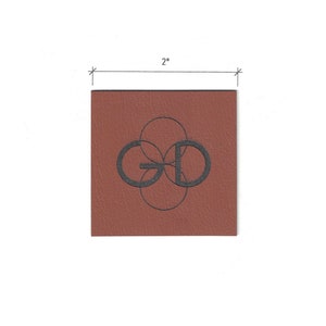 Peut inclure: Un patch en cuir carré de 5 cm de côté avec un fond brun et un logo noir en relief. Le logo est un "GD" stylisé avec des cercles qui se chevauchent.
