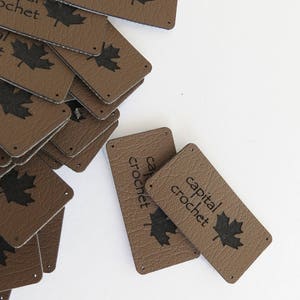 Puede incluir: Etiquetas de cuero marrón con el texto "capital crochet" y un diseño de hoja de arce negra. Las etiquetas son rectangulares con esquinas redondeadas y pequeños agujeros para coser. Un montón de etiquetas esparcidas sobre una superficie blanca.