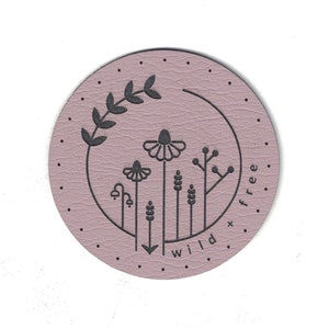 May include: A round, light purple leather patch with a black outline of wildflowers and a branch. The text "wild + free" is written in a black script font around the bottom of the patch.