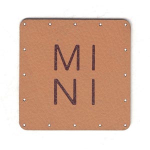 Può includere: Un'etichetta quadrata in pelle marrone chiaro con angoli arrotondati e la parola "MINI" stampata in marrone scuro. L'etichetta presenta piccoli fori distanziati uniformemente attorno al perimetro.
