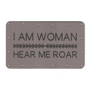 May include: A grey leather patch with the text "I AM WOMAN HEAR ME ROAR" in black lettering. A decorative line of leaves separates the two lines of text.