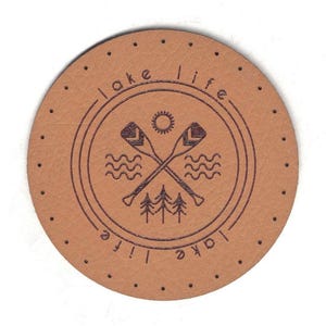 Può includere: Toppa rotonda in pelle marrone chiaro con la scritta "lake life" che circonda un disegno. Il disegno include remi incrociati, linee ondulate, un sole e pini. La toppa ha una serie di piccoli fori distanziati uniformemente attorno al bordo.