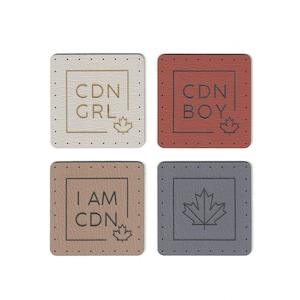 Può includere: Quattro toppe quadrate in pelle con diversi design. La prima toppa è bianca con il testo "CDN GRL" in lettere dorate e una foglia d'acero dorata. La seconda toppa è rossa con il testo "CDN BOY" in lettere nere e una foglia d'acero nera. La terza toppa è marrone con il testo "I AM CDN" in lettere nere e una foglia d'acero nera. La quarta toppa è grigia con una foglia d'acero nera.