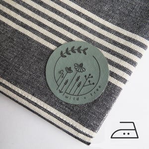 Op de afbeelding: Een close-up van een gevouwen, gestreepte stof in zwart en wit, met een cirkelvormige, saliegroene leren patch. De patch heeft een bloemenontwerp en de woorden "wild + free". Een strijkijzersymbool staat in de rechter benedenhoek.