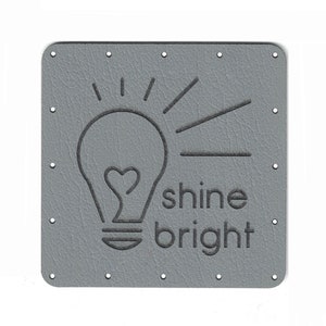 Puede incluir: Un parche cuadrado de cuero gris con esquinas redondeadas. Presenta un diseño de bombilla con un filamento en forma de corazón y las palabras "shine bright" en relieve en un gris más oscuro. Pequeños agujeros están espaciados uniformemente alrededor de los bordes.