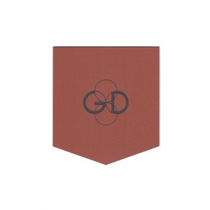 May include: A terracotta-colored leather bookmark with a geometric design and the letters "GD" in a circular pattern. The bookmark has a unique, pointed bottom edge and a textured surface.