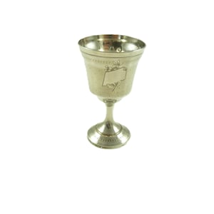 May include: A silver-colored goblet with a flared rim and a slender stem. The cup features a decorative band near the top and a blank rectangular cartouche with floral accents. The goblet is set against a white background.
