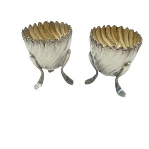 May include: Two silver-colored egg cups with a fluted design and three curved legs. The interior of each cup has a gold-colored finish. The egg cups are set against a white background.
