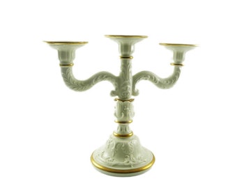May include: A white ceramic candelabra with three arms, adorned with gold trim. The base and arms feature ornate, embossed floral designs. The candelabra is a decorative piece for home decor.
