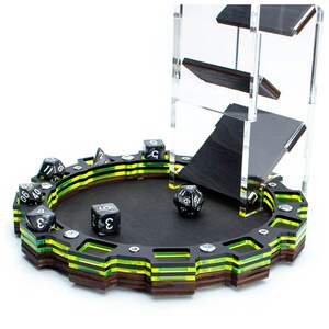 Op de afbeelding: Een zwarte en groene tafelspel dobbelsteentoren met een zwarte en gele ronde dobbelsteenrollade. De toren heeft drie niveaus en de lade heeft een zwart oppervlak met een gele en groene rand. Er liggen verschillende zwarte dobbelstenen met witte cijfers op de lade.