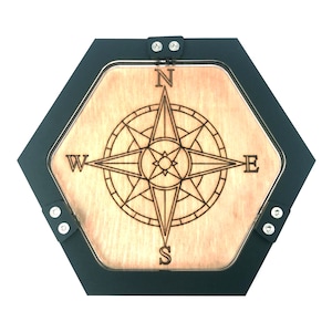 May include: A wooden compass rose design is laser-etched onto a light-colored wood surface, set within a black hexagonal frame. The compass points North, South, East, and West. The frame is secured with small silver rivets.