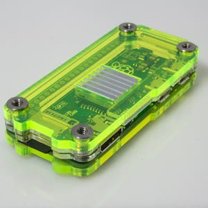 Puede incluir: Una carcasa de plástico verde para una computadora Raspberry Pi. La carcasa tiene una parte superior transparente y un disipador de calor plateado.