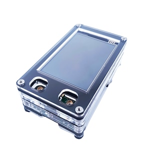 May include: A clear plastic case with a black frame and a small LCD screen. The case has two small holes on the top and a small circuit board visible inside.